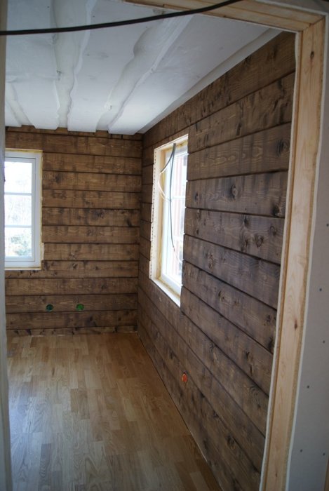 Interiör av ett rum med mörkbruna träpanelväggar, fönster och parkettgolv.