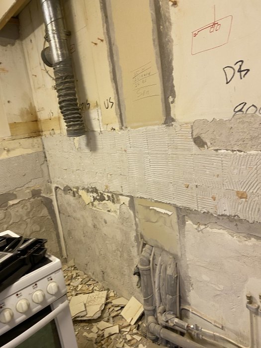 Ojämn vägg med delvis borttaget kakel och spackel, synlig rörledning och elledningar, vid en köksspis.