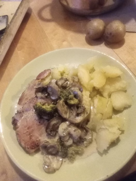 En tallrik med kassler, kokt potatis, champinjoner och broccoli i gräddsås.