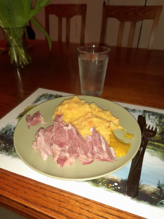 En tallrik med skivad skinka och mos på ett matbord, glas med vatten och envas i bakgrunden.