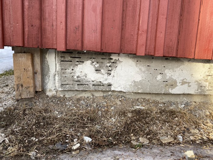 Del av rött trähus som vilar på en skadad betonggrund med synliga hålstenar och marken runtomkring.