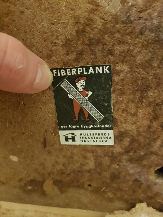 Etikett för "FIBERPLANK" från Hultsfreds Industrierna på en brunfiberplatta, fotograferat nära med en fingerpekning.