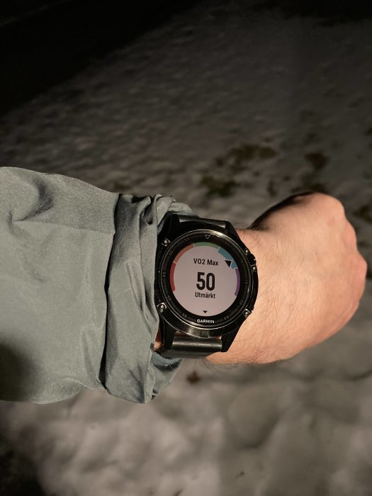 En handled som bär en Garmin sportklocka visar VO2 Max 50, med snötäckt mark i bakgrunden.