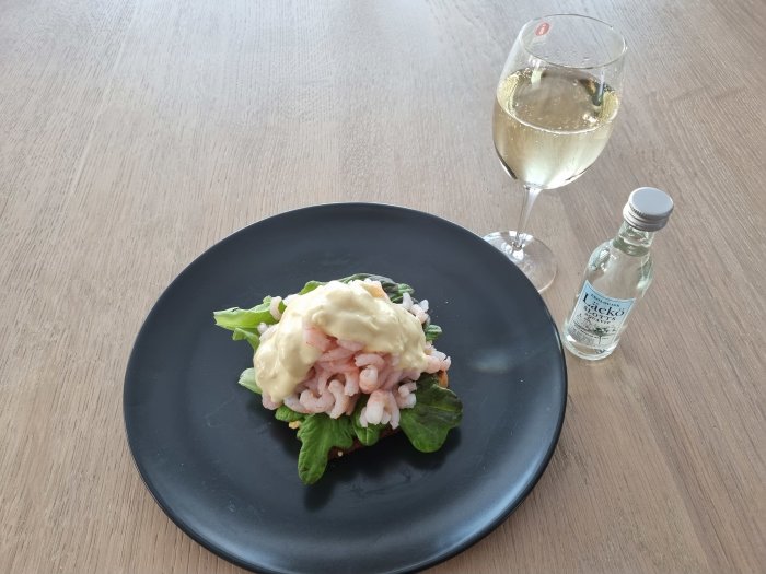 Räkmacka på svart tallrik med ett glas vin och en liten vodkaflaska på ett träbord.