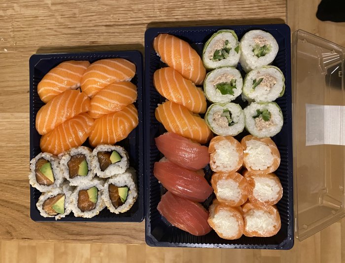 Fyra olika sorters sushi i förpackningar på träbord, inklusive lax nigiri och maki rullar.