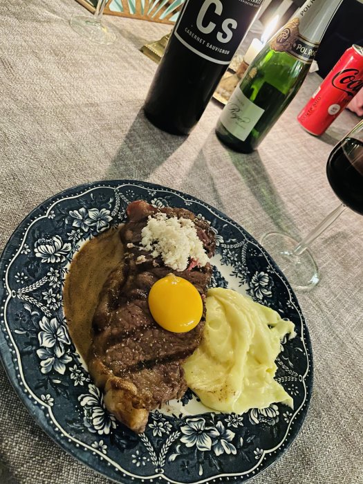 Ryggbiff med äggula, pepparrot och mandelpotatispuré på mönstrad tallrik, flankerad av vinflaskor och ett glas vin.