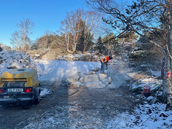 Person använder tryckluftsdriven bergborr på snötäckt mark med berg och träd i bakgrunden.