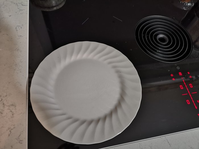Tom vit tallrik på svart spishäll med röda indikatorljus, tom för matlagning.