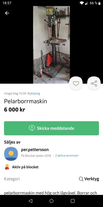 Gammal pelarborrmaskin i ett verkstadsrum med borrkronor upphängda på väggen och en röd skyddsduk på golvet.