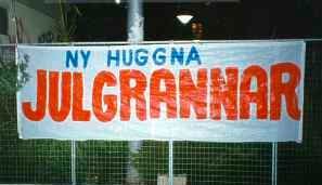Banner med texten "NY HUGGNA JULGRANNAR" uppställd utomhus på kvällen.