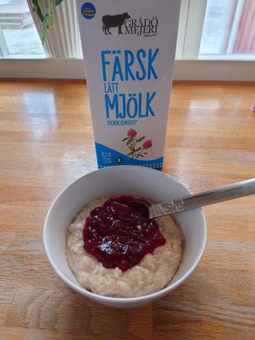 Frukost med gröt toppad med sylt och en mjölkpaket på ett köksbord framför ett fönster.