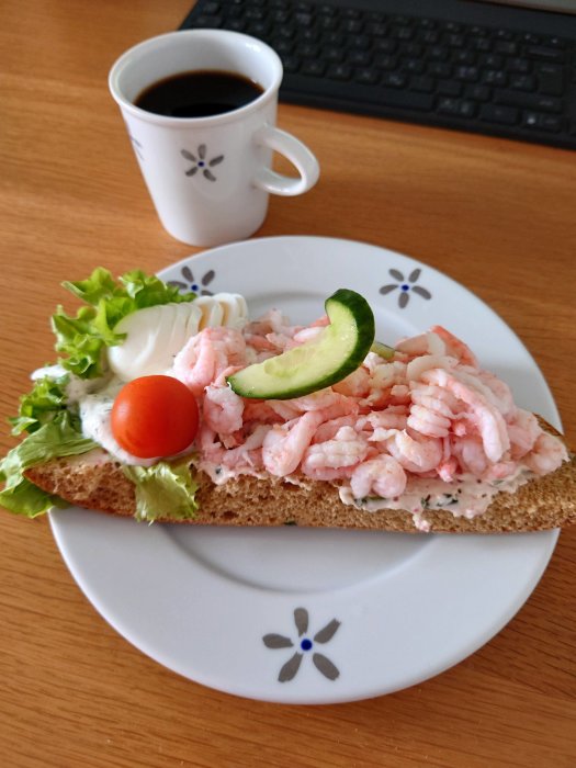 Räkmacka med sallad och majonnäs på tallrik bredvid kaffekopp och tangentbord.
