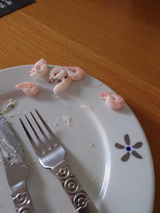 Nästan tomt vitt tallrik med några tillskurna räkor, en kniv och en gaffel på ett träbord.