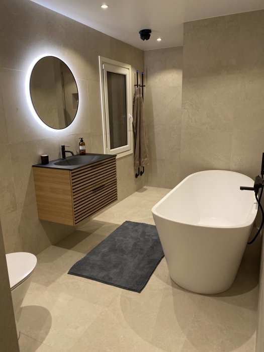Renoverat badrum med fristående badkar, rund spegel med belysning, träkommod och handdukar.