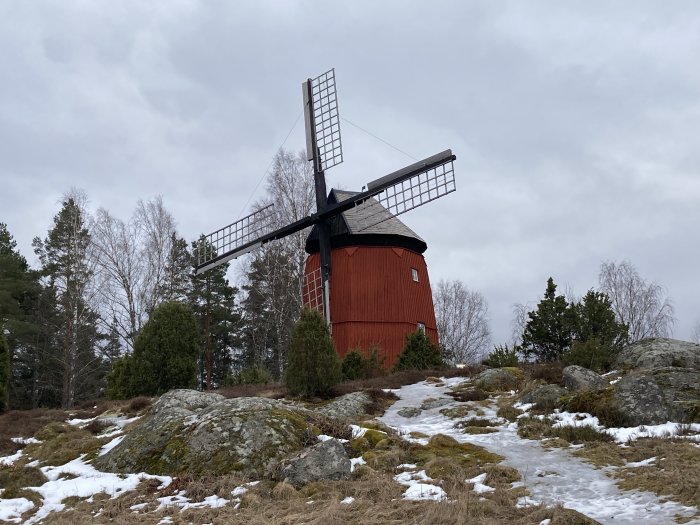 Röd väderkvarn på en kulle med smältande snö och träd i bakgrunden.