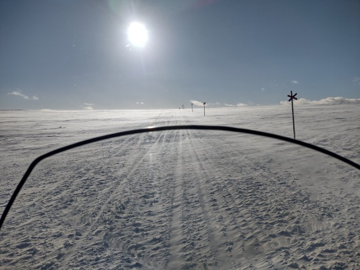 Solsken över snötäckt landskap med skidspår och ledstänger, sett genom skidglasögon.