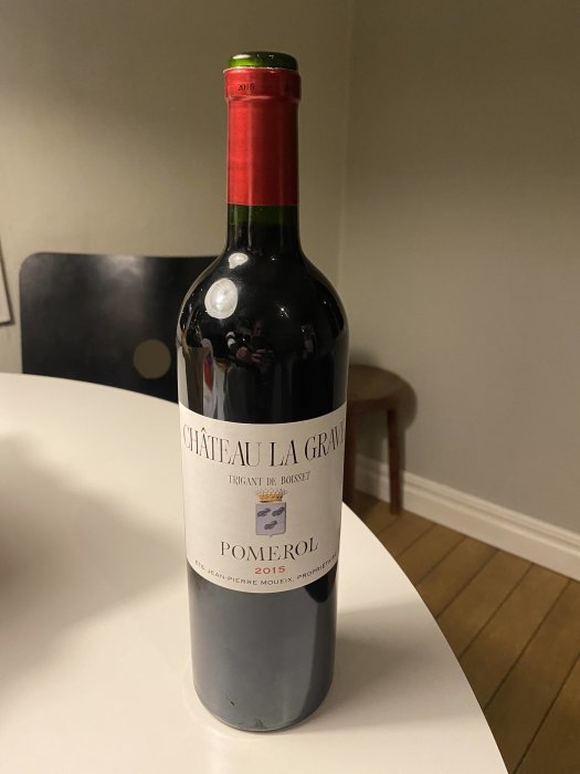 Flaska Château La Grave Pomerol från 2015 placerad på ett vitt bord mot en suddig bakgrund.