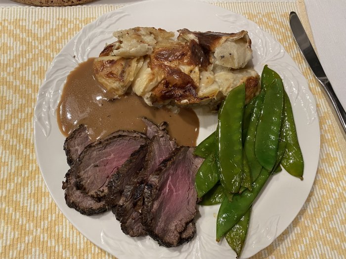 Skivad rostbiff med sås, potatisgratäng och brynta sockerärtor på en tallrik.