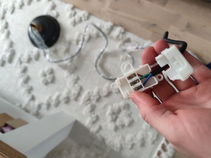 En hand som håller en elektrisk anslutning med lösa kablar mot en suddig bakgrund av en matta och lampa.