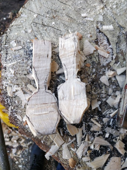 Två grovt utskurna träfigurer på en stubbe omgiven av spån och bark.