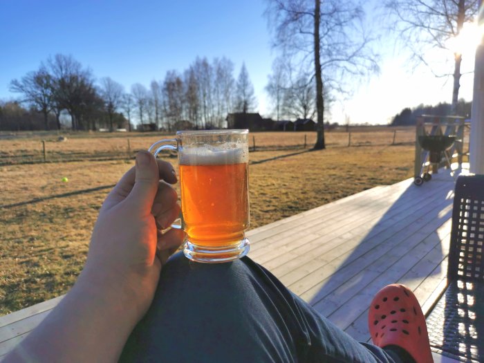 Hand håller ett glas öl på en solig veranda med en grill och landskap i bakgrunden.