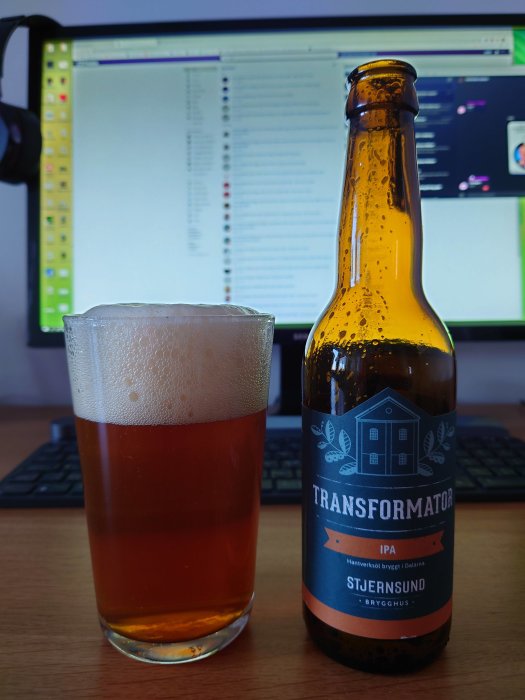 Ett glas öl bredvid en ölflaska med etiketten "Transformator IPA" framför en datorskärm.