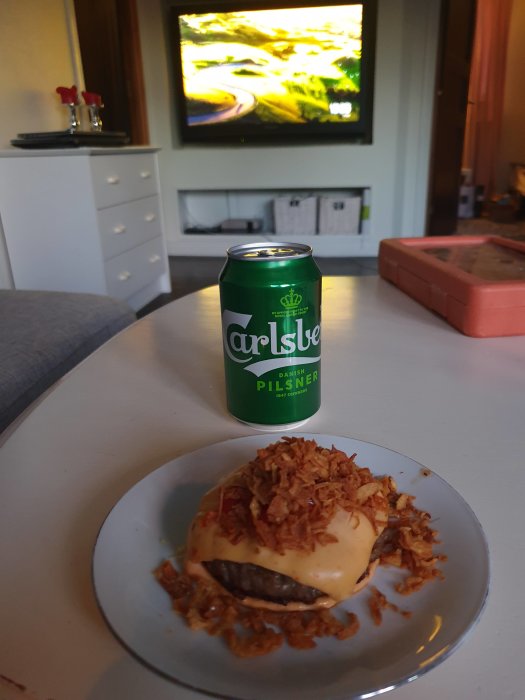 Hamburgare med ost och rostad lök på tallrik, burk öl bredvid, med suddig TV i bakgrunden.