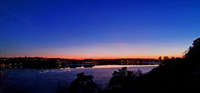 Solnedgång över en stad med silhuetter av byggnader och spegling i en flod.