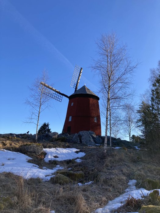 Röd väderkvarn på klippig mark med snöfläckar under en klarblå himmel.