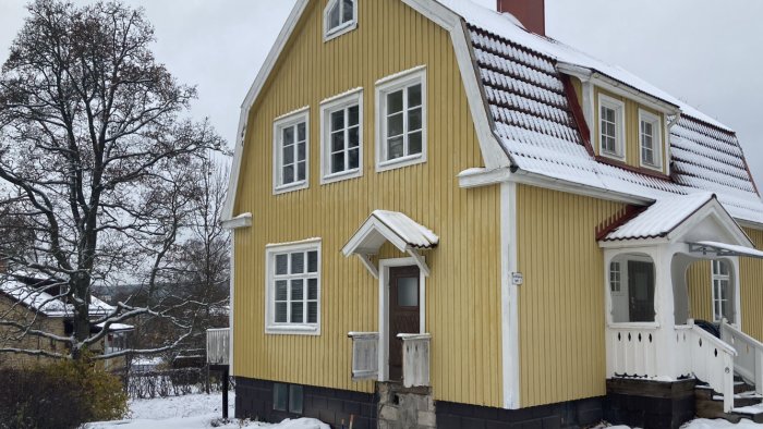 Gult traditionellt trähus med snötäckt tak, balkong och utan synlig tillbyggnad.