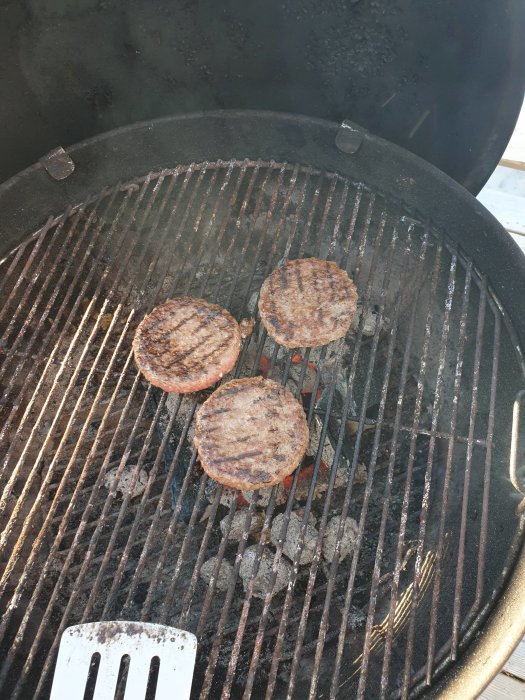 Tre hamburgare som grillas på en kolgrill med en stekspade i förgrunden.