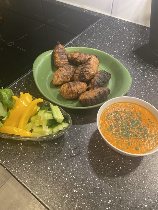 Grillade biffar på ett grönt fat med mojosås och skivad gurka samt gul paprika på köksbänk.