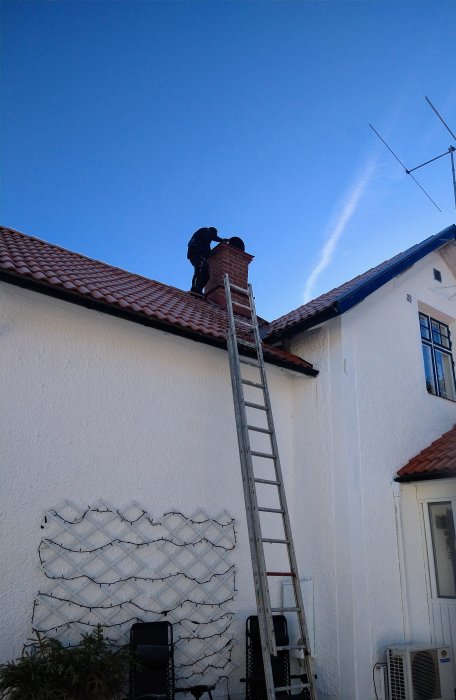 Sotare på stege vid skorstenen på ett rött tegeltak mot en klarblå himmel.