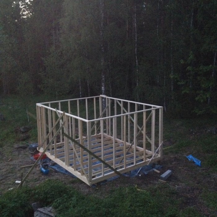 Grundstrukturen av ett träbygge i skymningen med skogsbakgrund och byggmaterial på marken.