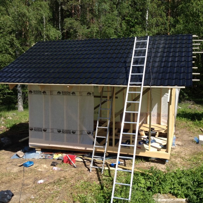 Halvfärdig träkonstruktion av ett litet hus med svart tak och stege, omgivet av verktyg och byggmaterial.