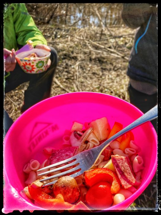 En skål med pastasallad med makaroner, salami, paprika, tomat och ost under en utomhusvandring.