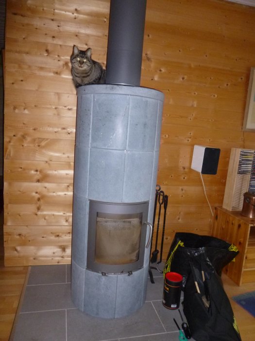 Kamin av sten omgiven av liggande pärlspontvägg med en katt på toppen, verktyg och eldstadsredskap.