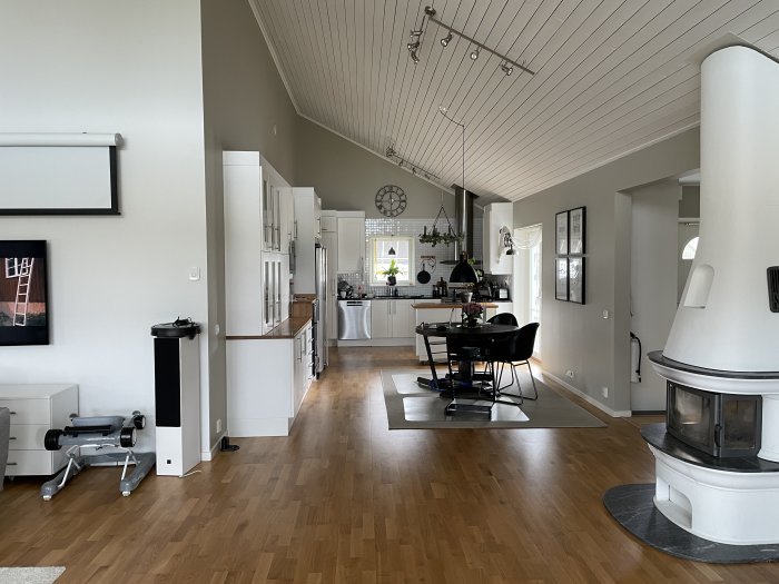 Öppen planlösning med kök i bakgrunden, matsalsbord och centralt placerad öppen spis, trägolv och vitmålade väggar.