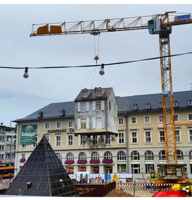 Byggnad som lyfts av en kran mitt i en stadsmiljö, med byggnadsställningar i bakgrunden.