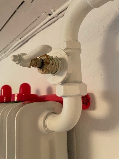 En gammal radiatorventil med synliga rost och kalkavlagringar som läcker, monterad på en vitmålad radiator.