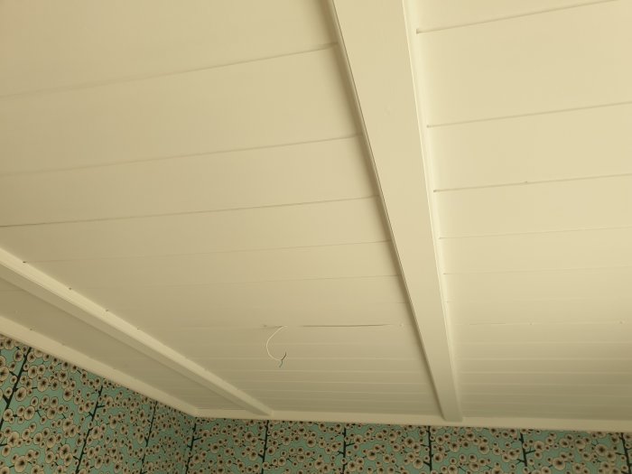 Vitlackerat träpaneltak med synlig spricka och färgskrap nära taklist mot mönstrat golv.