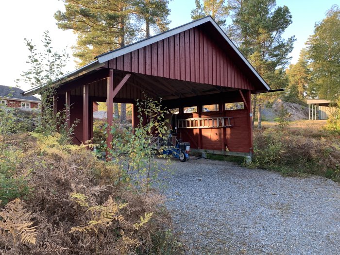 En röd öppen carport på betongplintar med tak, omgiven av grönska och grusväg, under ombyggnadsövervägande.