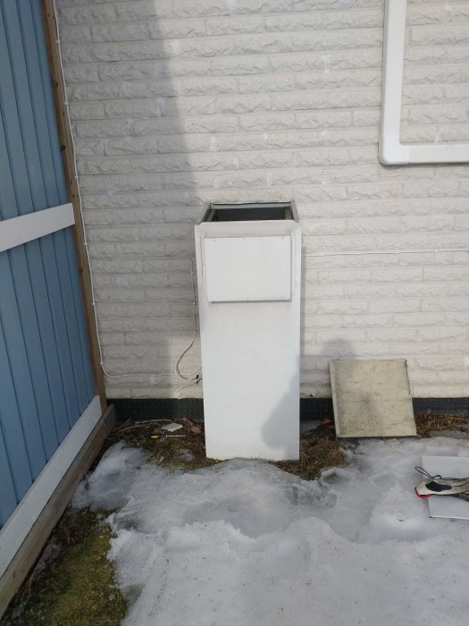 Vit, okänd utomhusinstallation vid husvägg med öppet lock och synliga rörledningar i snöigt landskap.