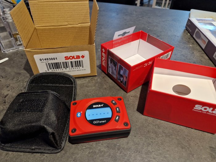 SOLA digital inclinometer with a display, alongside its packaging boxes and a protective carrying pouch on a table.