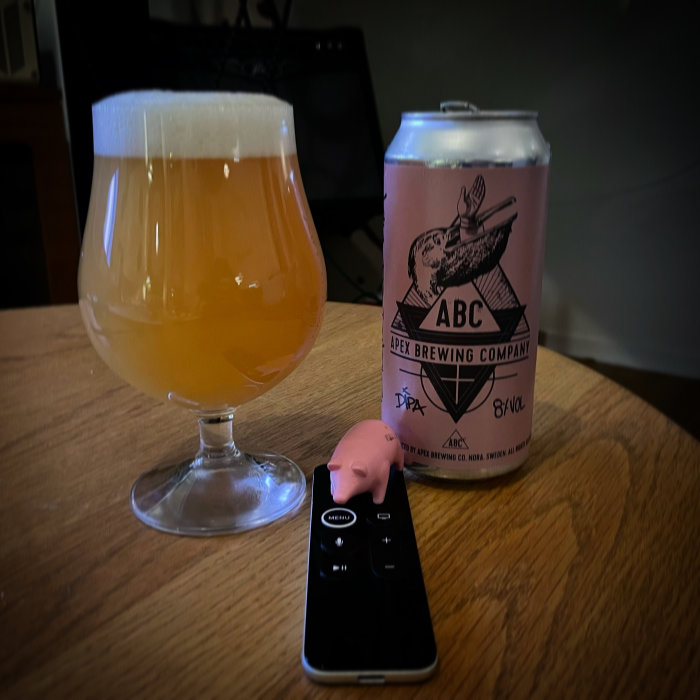 Ölglas med skum på ett bord, en Apex Brewing ölburk och en fjärrkontroll vid sidan.