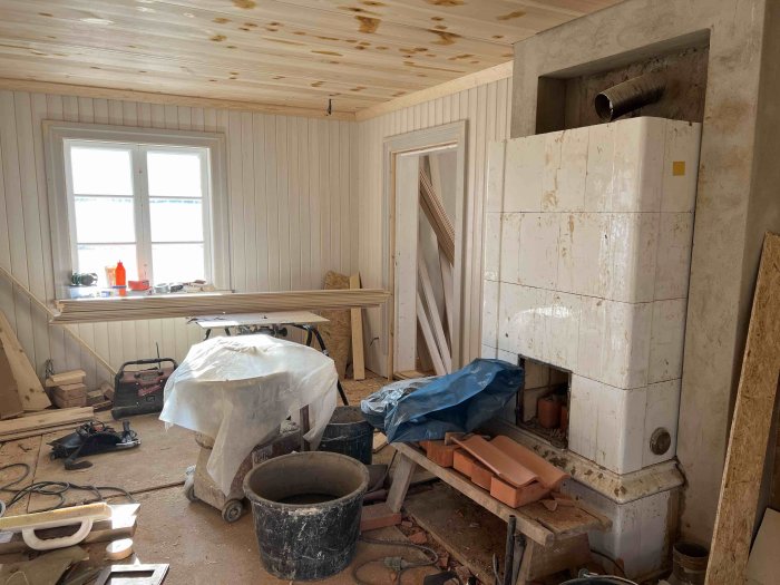 Rum under renovering med en oavslutad kakelugn omgiven av byggmaterial och verktyg.