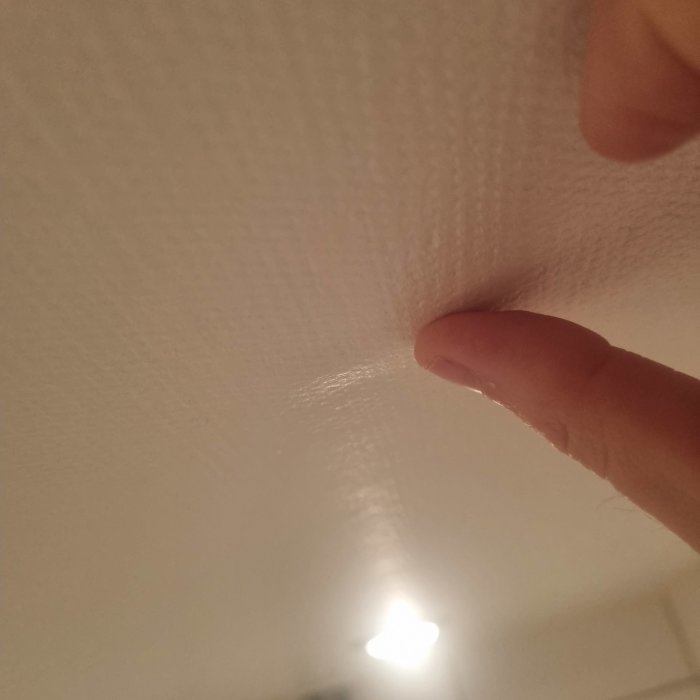 Finger pekar på en bubbla i väven i badrumstaket under en lampa, med funderingar om målning eller spackling.