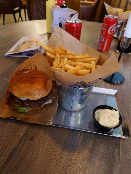 Hamburgare med pommes frites på restaurangbord, med läskburkar och meny i bakgrunden.