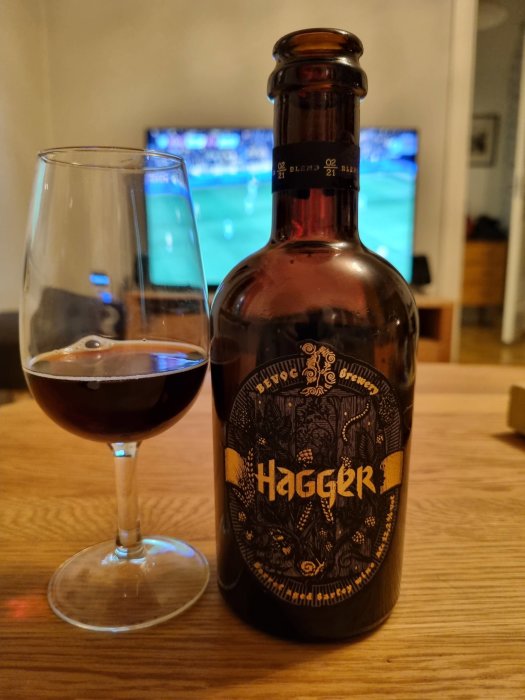 Flaska och glas med barley wine framför suddig fotbollsmatch på TV.