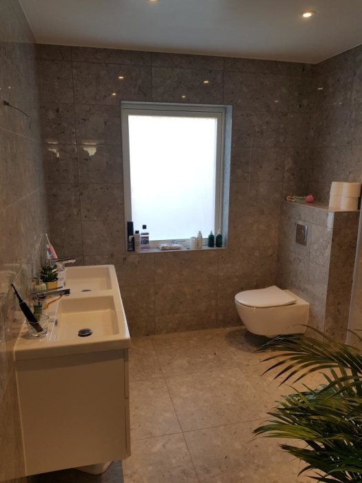 Modernt badrum med marmorkaklade väggar, integrerad handfat, toalett och produkter längs fönsterkarmen.